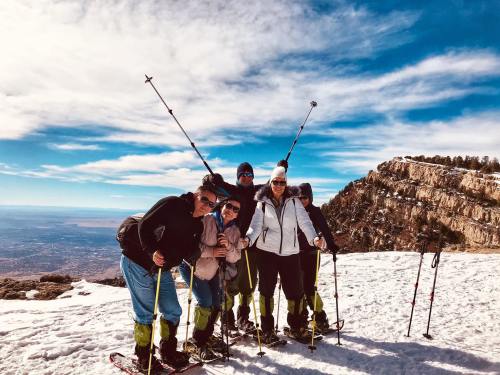 A group of six people poses on a snowy mountain, smiling with ski poles against a backdrop of blue sky and rocky terrain.