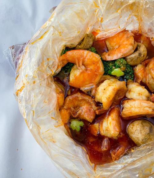 A close-up of a seafood dish in a plastic bag, featuring shrimp, broccoli, and mushrooms in a spicy sauce.