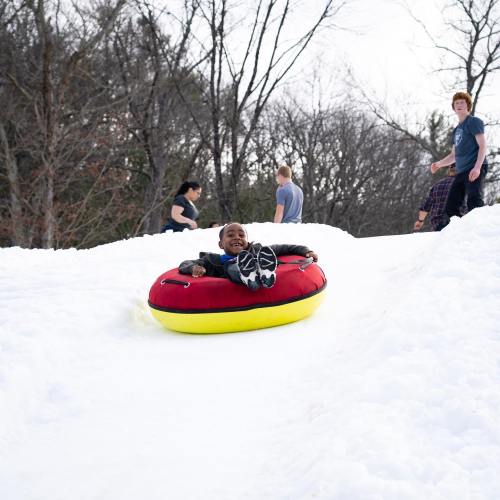 A child joyfully sleds down a snowy hill on a red and yellow tube, with people playing in the background.