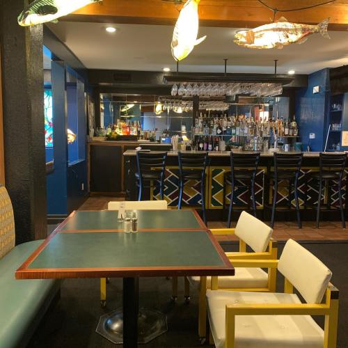 A cozy restaurant interior featuring a bar, colorful decor, and a table with white chairs. Fish-shaped lights hang above.