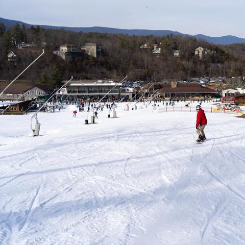 A snowboarder glides down a snowy slope with a ski resort and people in the background enjoying winter activities.