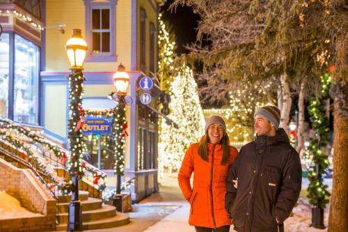 A couple walks hand in hand through a festive, snow-lit street adorned with holiday lights and decorations.