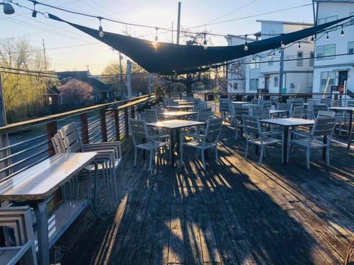 Outdoor dining area with wooden deck, tables, and chairs, illuminated by sunlight and shaded by a large canopy.