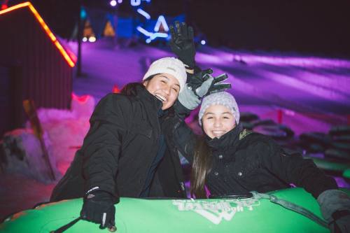 Two smiling individuals in winter attire pose with their hands raised, surrounded by a colorful, illuminated snow tubing area.