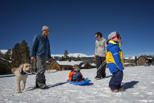 A family enjoys a snowy day, with a dog, sledding, and mountains in the background under a clear blue sky.