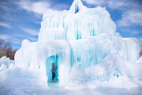 A person stands at the entrance of a large, icy structure with blue-tinted icicles under a bright blue sky.
