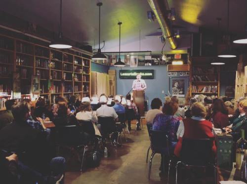 A speaker stands at a podium in a cozy bookstore, addressing an audience seated in rows. Bookshelves line the walls.