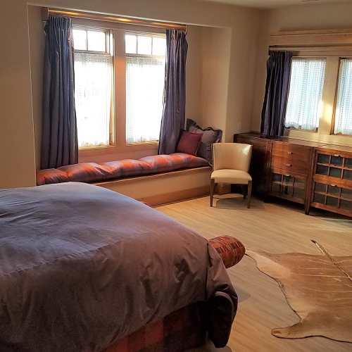 Cozy bedroom with a bed, window seat, chair, and wooden dresser, featuring natural light and warm decor.