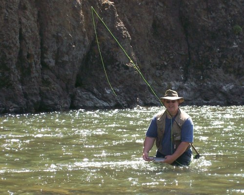 A person fishing in a river, wearing a hat and vest, with a fishing rod in hand and rocky cliffs in the background.