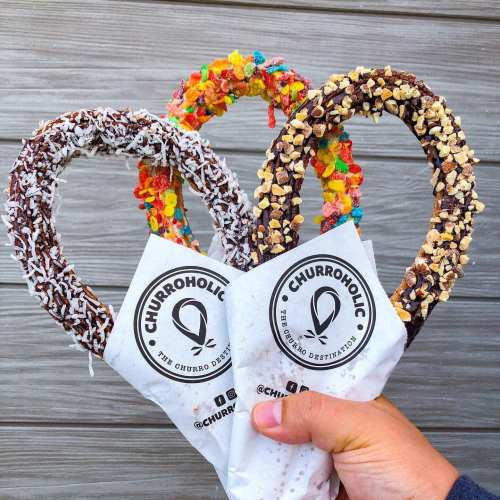 A hand holds three colorful churros coated in toppings, wrapped in paper with the logo "Churroholic."