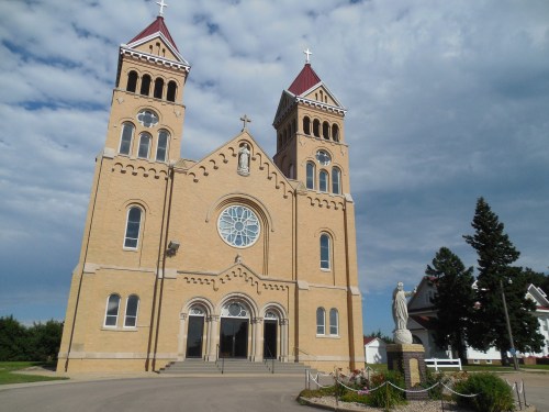 A large brick church with two towers, a rose window, and a statue in front, set against a cloudy sky.