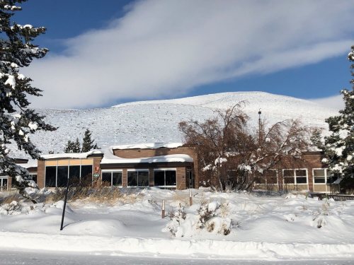 A snow-covered building with large windows, surrounded by snowdrifts and trees, against a backdrop of a snowy hill.