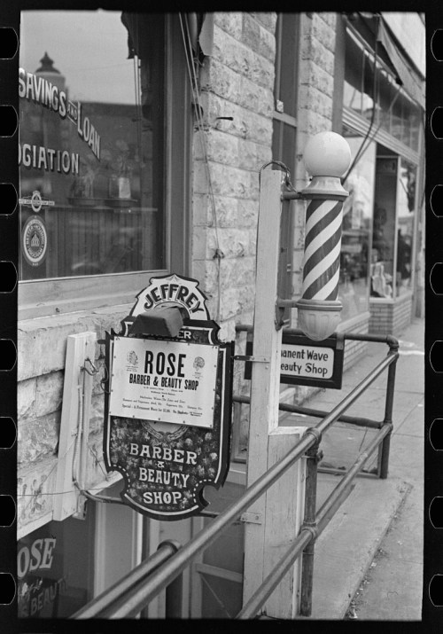 Black and white photo of a barber and beauty shop sign outside a building, featuring a classic barber pole.