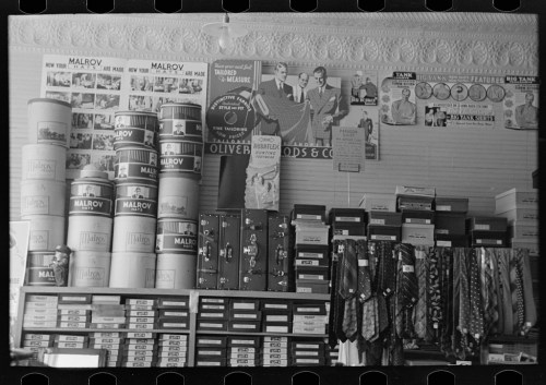 A vintage store display featuring stacked cans, boxes, and ties, with posters on the wall advertising various products.