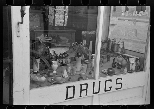 A vintage storefront window displaying various items, with the word "DRUGS" prominently painted on the glass.