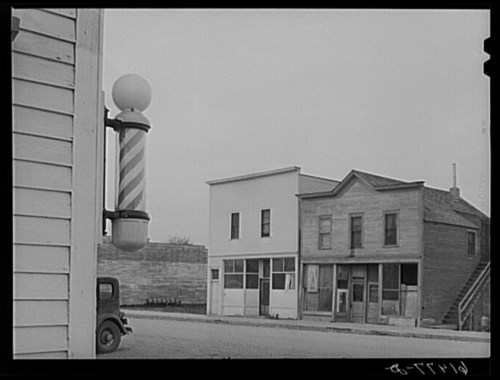 A vintage street scene featuring a barber pole, old buildings, and a parked car in a small town.
