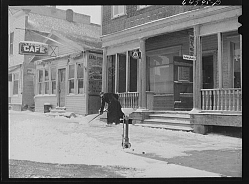 A person shovels snow in front of a café and shops on a snowy street.