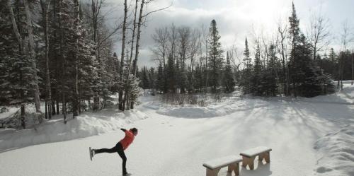 A person ice skating on a snowy pond surrounded by trees, with a cloudy sky and sunlight peeking through.