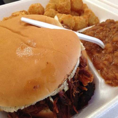 A close-up of a pulled pork sandwich with a side of tater tots and baked beans on a takeout tray.