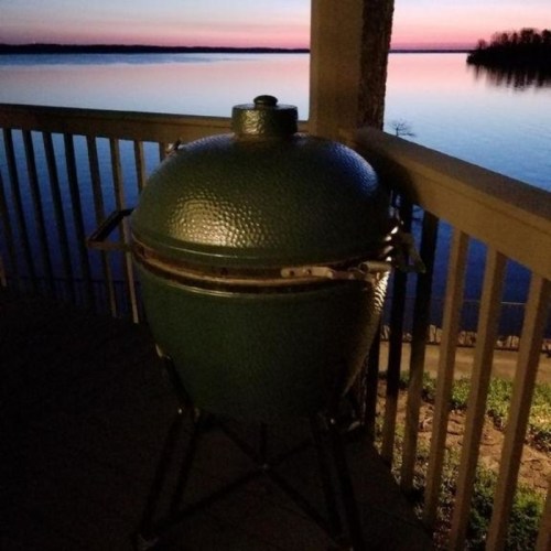 A green grill sits on a deck overlooking a calm lake at sunset, with vibrant colors reflecting on the water.