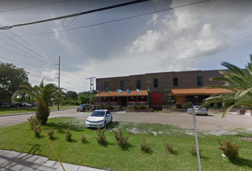 A brick building with flags, surrounded by palm trees and grass, with a parked white car in front.