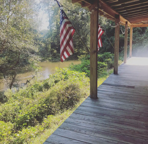 A wooden porch with American flags, overlooking a lush green area and a calm river in the background.