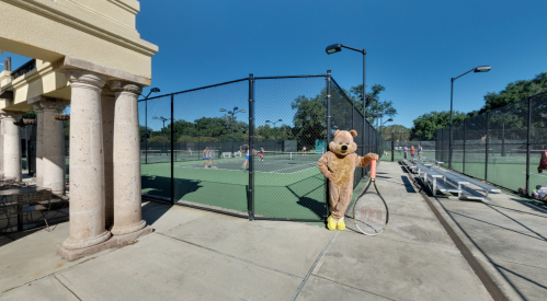 A bear mascot stands by a tennis court, holding a racket, with players visible in the background under a clear blue sky.