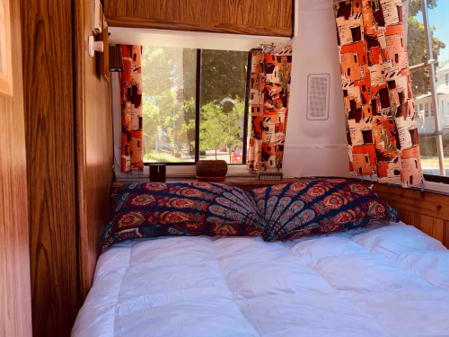 Cozy camper interior featuring a bed with colorful pillows and patterned curtains, illuminated by natural light.