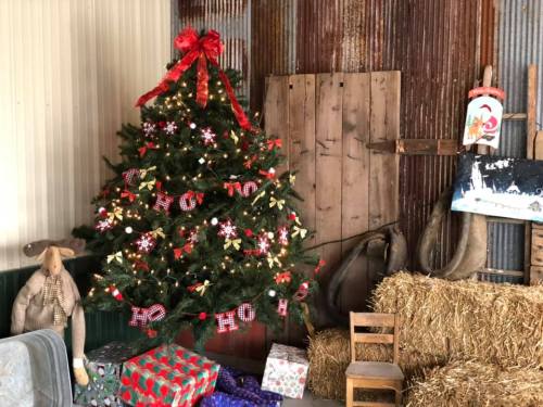 A decorated Christmas tree with gifts and rustic decor in a barn setting, featuring hay bales and wooden elements.