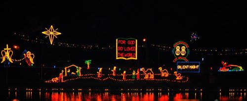 Colorful holiday lights display featuring a nativity scene, stars, and the text "For God so loved the world" by a waterway.