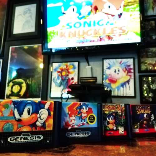 A collection of Sega Genesis game cartridges with a Sonic the Hedgehog theme, displayed in front of a TV showing Sonic.