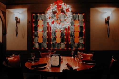 A cozy restaurant table set for dinner, featuring elegant glassware and a festive wreath on a stained glass window.