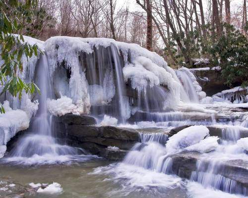 A frozen waterfall cascading over rocks, surrounded by icy formations and bare trees in a winter landscape.