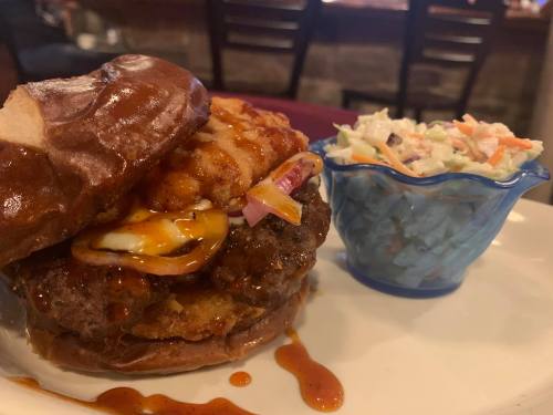A close-up of a burger with a pretzel bun, topped with cheese and onion, served with a side of coleslaw.