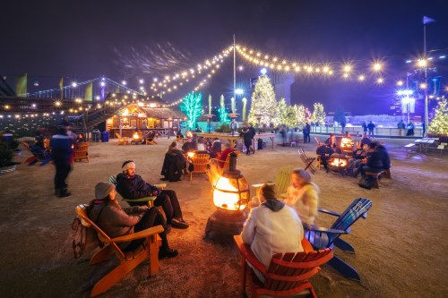 A festive outdoor scene with people gathered around fire pits, string lights, and decorated trees at night.
