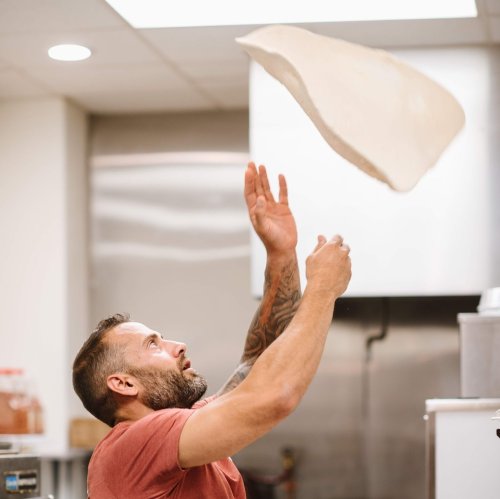 A man with tattoos tosses pizza dough in a kitchen, focused on the airborne dough above him.