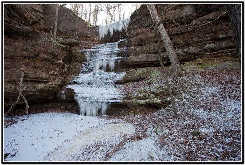 A frozen waterfall cascades down rocky cliffs, surrounded by trees and a snowy ground.