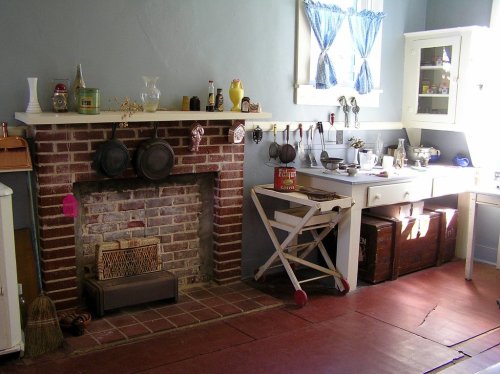 A vintage kitchen featuring a brick fireplace, wooden cabinets, and various kitchen utensils on a countertop.