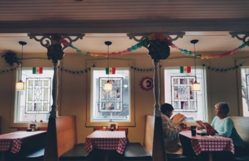 Two women sit at a table in a cozy restaurant, reading menus under colorful decorations and stained glass windows.