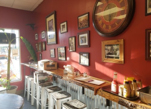 Cozy café interior with red walls, framed photos, a wooden counter, and condiments on display. Sunlight streams in.