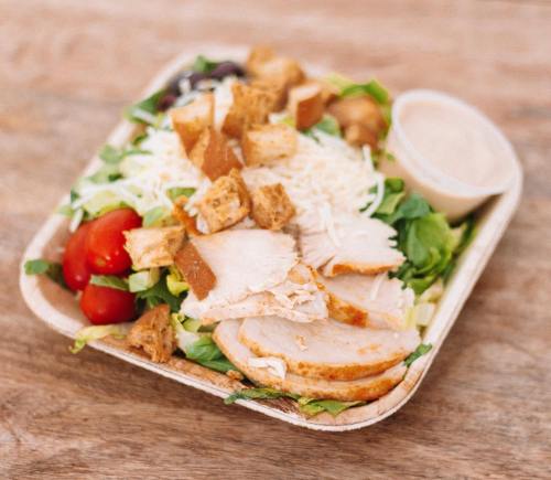 A salad with sliced chicken, lettuce, tomatoes, croutons, cheese, and a side of dressing in a compostable container.