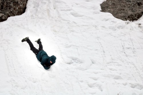 A person in a teal jacket lies on their stomach in a snowy landscape, surrounded by rocky terrain.