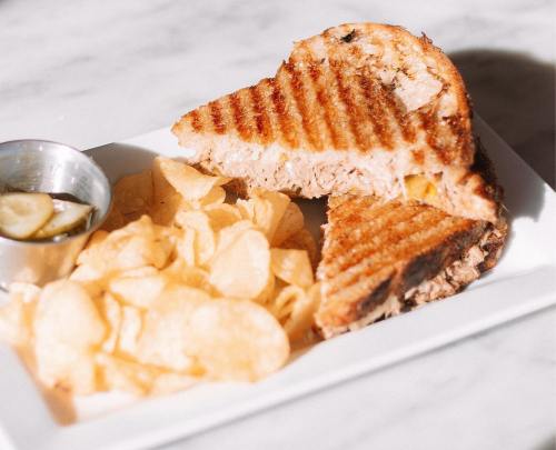 A grilled sandwich cut in half, served with a side of potato chips and a small dish of dipping sauce.
