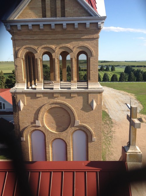 View of a brick church tower with arched windows, set against a green landscape and blue sky.