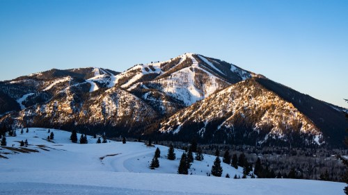 Snow-covered mountains under a clear blue sky, with sunlight illuminating the peaks and surrounding trees.