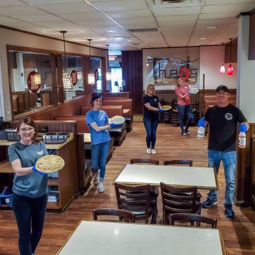 Five restaurant staff members hold pizzas and cleaning supplies in a spacious dining area, smiling at the camera.