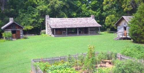 A scenic view of two rustic cabins surrounded by greenery and a garden in a peaceful outdoor setting.