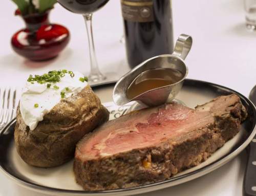 A plate with sliced prime rib, a baked potato topped with sour cream and chives, and a gravy boat, with wine in the background.