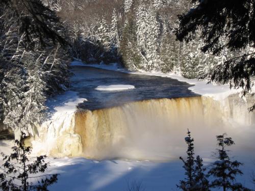 A snowy landscape featuring a waterfall cascading into a river, surrounded by evergreen trees and winter scenery.