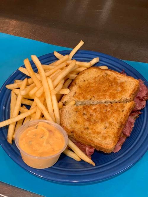 A toasted sandwich with meat, served with a side of fries and a small container of dipping sauce on a blue plate.